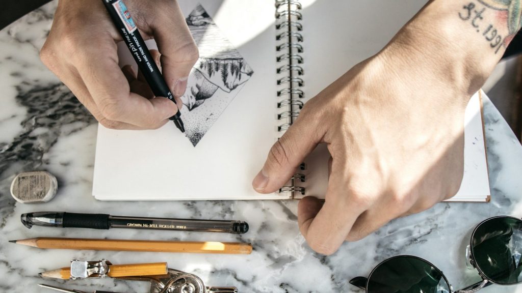 Hands sketching a design in a notebook, surrounded by pens and pencils.